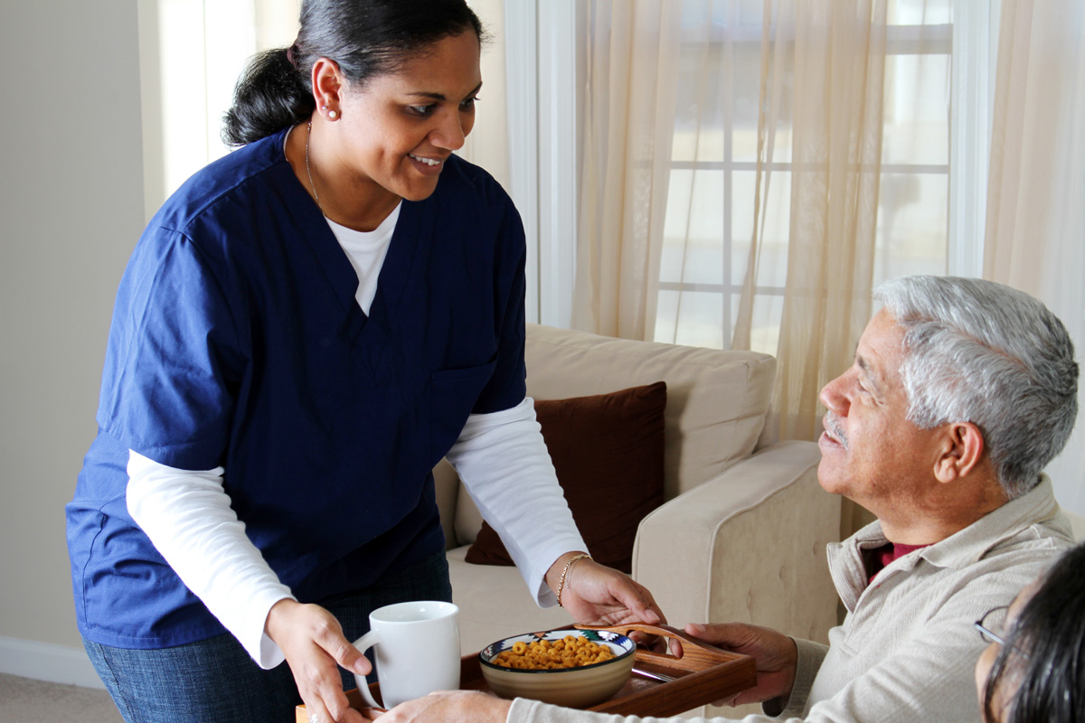 An in-home caregiver giving an elderly man a tray of food in El Paso.